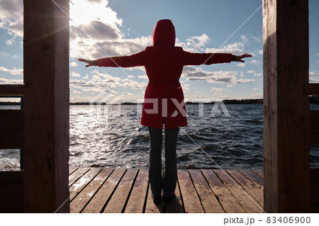 A woman in a Red Jacket Sits at a Table on a Wooden Pier and Looks at the Water, Clouds and Trees with Yellow Foliage. Autumn Landscape. A woman in a Red Jacket Sits at a Table on a Wooden Pier and Looks at the Water, Clouds and Trees with Yellow Foliage. Autumn Landscape. 83406900
