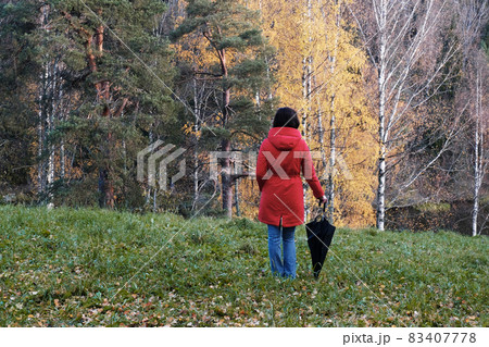 A Woman in a Red Jacket with an Umbrella Walks through the Autumn Park among the Yellow Fallen Leaves. A Woman in a Red Jacket with an Umbrella Walks through the Autumn Park among the Yellow Fallen Leaves. 83407778