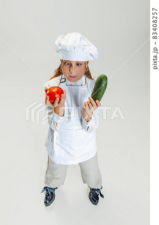 High angle view of little cute girl in white cook uniform and huge chef's hat posing isolated on white studio background. 83408257