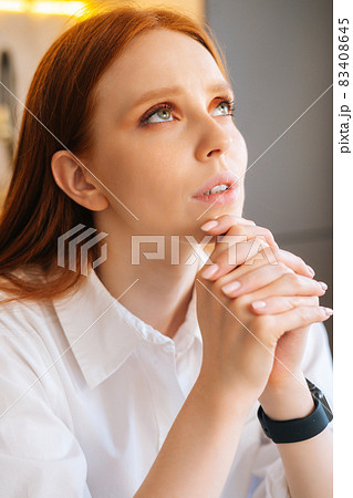 Close-up face of sad young woman praying holding folded hands in front of face at home, looking up. 83408645