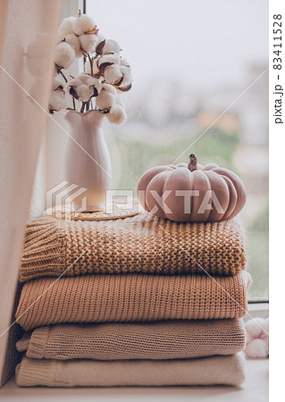 A stack of warm knitted sweaters on the windowsill with pumpkin and a vase of cotton plant on a rainy day, in pastel shades. 83411528