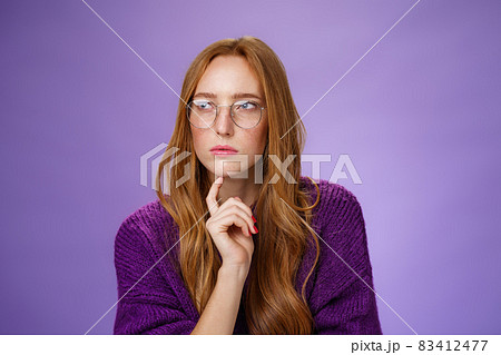 Girlfriend solving puzzle like detective making assumptions focusing on problem squinting looking left determined and thoughtful wearing glasses touching jaw while thinking over purple background 83412477