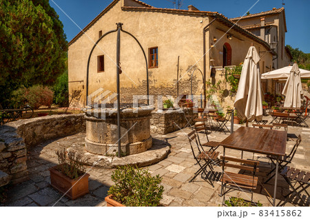 Bagno Vignoni, Tuscany, Italy. August 2020. In the center of the village a restaurant with outdoor tables and umbrellas, an ancient well gives the context a lot of charm. Bagno Vignoni, Tuscany, Italy. August 2020. In the center of the village a restaurant with outdoor tables and umbrellas, an ancient well gives the context a lot of charm. 83415862