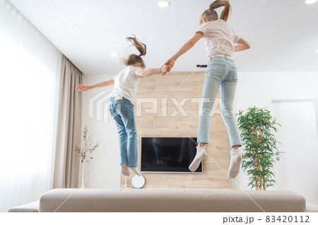 Two sisters jumping on couch in living room having fun Two sisters jumping on couch in living room having fun 83420112