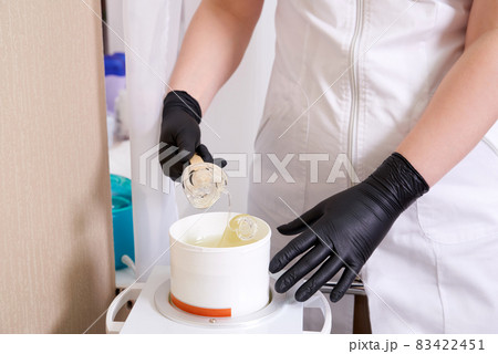 Wax heater with hot yellow wax on wooden spatula in human hand. Hot wax for depilation. Salon equipment Wax heater with hot yellow wax on wooden spatula in human hand. Hot wax for depilation. Salon equipment 83422451