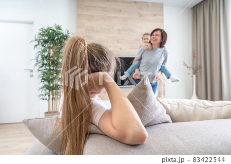 Preteen sad girl sitting on couch while mother having fun with younger sister, jealousy concept Preteen sad girl sitting on couch while mother having fun with younger sister, jealousy concept 83422544