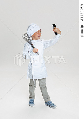 Close-up portrait of little happy boy in white cook uniform and huge chef's hat using phone isolated on white studio background. Close-up portrait of little happy boy in white cook uniform and huge chef's hat using phone isolated on white studio background. 83432059