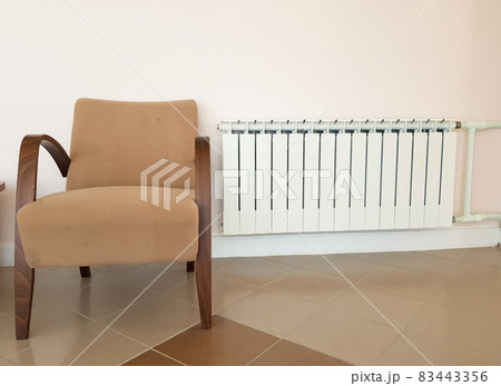 A modern aluminum white heating radiator and a fashionable brown armchair on the background of a wall in a residential area A modern aluminum white heating radiator and a fashionable brown armchair on the background of a wall in a residential area 83443356