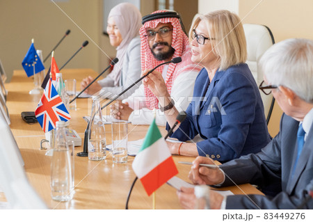 Mature female politician in eyeglasses sitting among multi-ethnic colleagues and speaking into microphone at conference 83449276