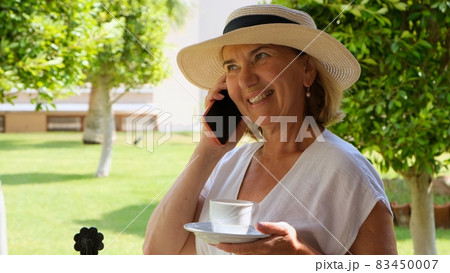 blonde senora woman, 50-55 years old, in a hat and a white dress, talks using a smartphone and holds 83450007