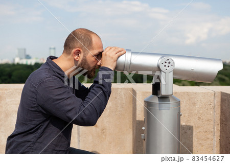 Man tourist standing on building rooftop looking through binocular telescope Man tourist standing on building rooftop looking through binocular telescope 83454627