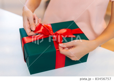 Close-up top view of unrecognizable female hands wrapping Christmas gift box tying red ribbon and decorating beautiful bow, closeup. 83456757