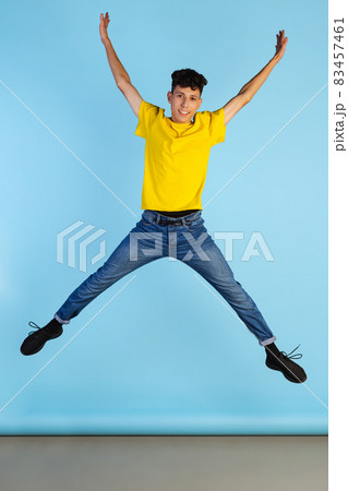 Full-length portrait of young boy in yellow T-shirt jumping isolated over blue background. Happiness Full-length portrait of young boy in yellow T-shirt jumping isolated over blue background. Happiness 83457461