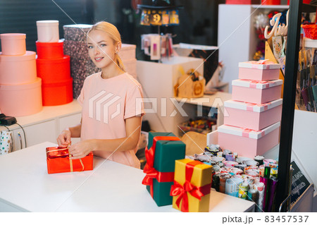 Attractive young saleswoman holding and using golden ribbon to tie bow for red wrapped gift box for Christmas present at counter of holiday shop. 83457537