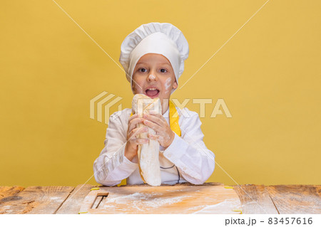One cute boy wearing in white cook, chef jacket and hat kneads dough isolated on yellow studio background. One cute boy wearing in white cook, chef jacket and hat kneads dough isolated on yellow studio background. 83457616