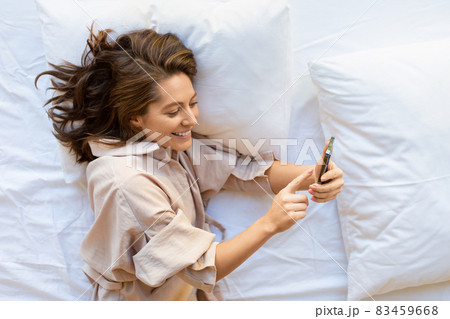 Top view of a smiling 35-years-old woman with smartphone in bed. 83459668