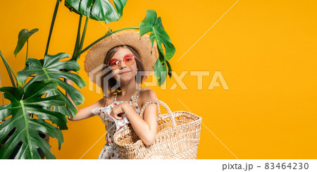 Pre-teen girl in straw hat, orange sunglasses holds straw handbag while stands near tropical plants Pre-teen girl in straw hat, orange sunglasses holds straw handbag while stands near tropical plants 83464230