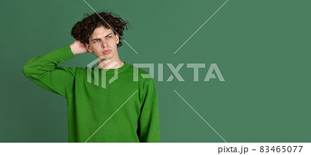 Cropped portrait of young man in green sweater with facial expression of misunderstanding isolated over dark green background Cropped portrait of young man in green sweater with facial expression of misunderstanding isolated over dark green background 83465077