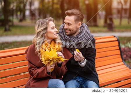 Embracing man found a small leaf happy couple sitting on the bench hugged in park wearing coats and scarfs collecting a bouquet of fallen leaves. Love story concept. Tinted image Embracing man found a small leaf happy couple sitting on the bench hugged in park wearing coats and scarfs collecting a bouquet of fallen leaves. Love story concept. Tinted image 83466202