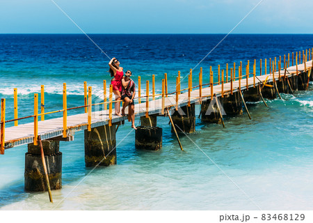 A happy couple on vacation strolling along a wooden pier above the tropical, turquoise ocean in Bali, Indonesia. Couple in romantic walk along tropical ocean pier. Lovers walk along the pier A happy couple on vacation strolling along a wooden pier above the tropical, turquoise ocean in Bali, Indonesia. Couple in romantic walk along tropical ocean pier. Lovers walk along the pier 83468129