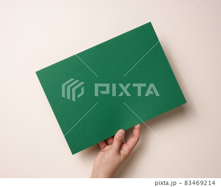 Female hand holding empty green paper on a beige background. Copy space 83469214