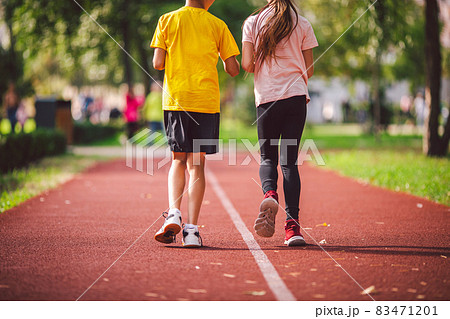 Topic is sports, physical development, athletics training for schoolchildren in physical education lessons. Close-up of legs two children boy and girl, running across red running stadium, rear view 83471201