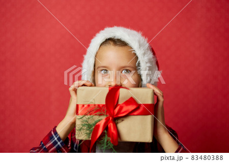 Cute little girl in santa hat holding Christmas gifts in front of her face 83480388