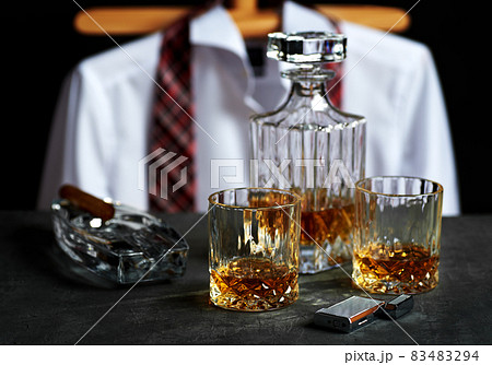 Bottle and two glasses of whiskey on a stone table top in a bar. Lighter and Cuban cigar with a white shirt and red tie. Bottle and two glasses of whiskey on a stone table top in a bar. Lighter and Cuban cigar with a white shirt and red tie. 83483294