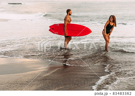 Beautiful couple have fun at the sea. Couple in love on the beach with a surfboard at sunset. Beautiful couple on the island of Bali. Couple of surfers walking on coast in Indonesia. Vacation at sea 83483362