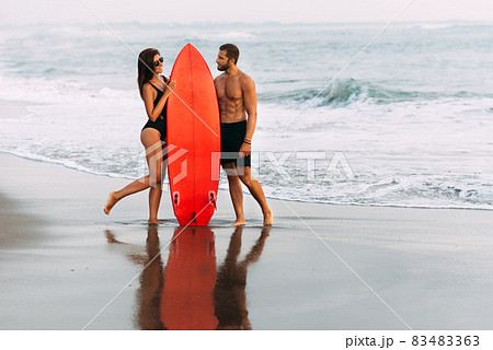 A couple in love on the beach with a surfboard. Beautiful couple on the island of Bali. Happy sporty couple kissing behind surf boards on the beach at sunset. The concept of a healthy lifestyle A couple in love on the beach with a surfboard. Beautiful couple on the island of Bali. Happy sporty couple kissing behind surf boards on the beach at sunset. The concept of a healthy lifestyle 83483363