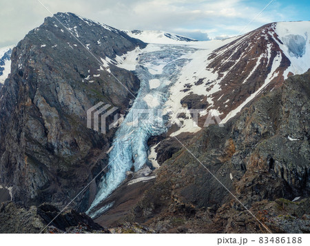 Inaccessible large glacier and great snowy mountain. Scenic highlands landscape with big cracked blue glacier with scratches among moraines on background of high snowy mountain peak and rock top. Inaccessible large glacier and great snowy mountain. Scenic highlands landscape with big cracked blue glacier with scratches among moraines on background of high snowy mountain peak and rock top. 83486188