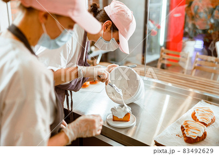 Confectioner trainee spreads cream onto sponge cake while colleague teaches her in bakery Confectioner trainee spreads cream onto sponge cake while colleague teaches her in bakery 83492629