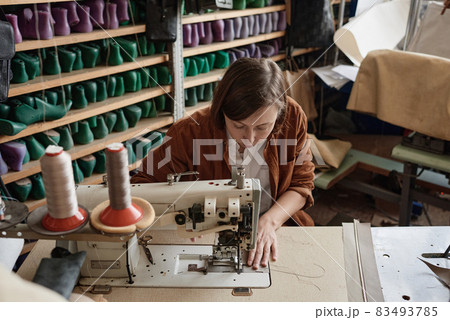 Shoemaker sitting at the table and using sewing machine in her work, she sewing new shoes Shoemaker sitting at the table and using sewing machine in her work, she sewing new shoes 83493785