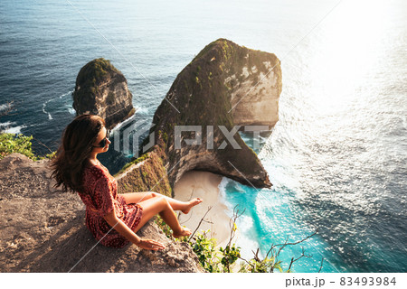 Beautiful girl sitting on a high rock and looking out to sea. Girl sits on the edge of cliff. The girl of the sea. Seascape. Girl at sunset. Sea tour. Nusa Penida Bali Indonesia 83493984