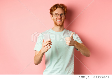 Happy young man with red messy hair, showing thumbs-up while using mobile phone, smiling and praising application, standing over pink background Happy young man with red messy hair, showing thumbs-up while using mobile phone, smiling and praising application, standing over pink background 83495821
