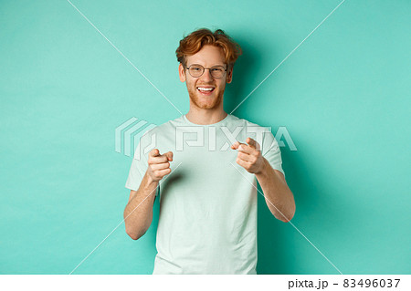 Handsome young man with ginger hair, wearing glasses and t-shirt, pointing finger at camera and smiling, choosing you, congratulating or praising, standing over mint background Handsome young man with ginger hair, wearing glasses and t-shirt, pointing finger at camera and smiling, choosing you, congratulating or praising, standing over mint background 83496037