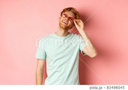 Tourism and vacation concept. Happy young man with red hair relaxing, wearing sunglasses and smiling carefree, standing over pink background 83496045