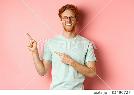 Portrait of cheerful young man with red hair, wearing glasses, pointing fingers at upper left corner and smiling, standing over pink background Portrait of cheerful young man with red hair, wearing glasses, pointing fingers at upper left corner and smiling, standing over pink background 83496727