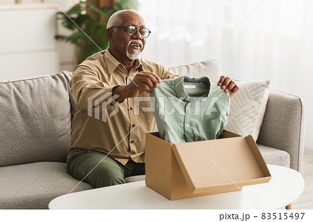 Senior Black Man Unpacking Box Looking At Shirt At Home 83515497