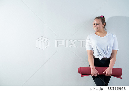 Young fat caucasian woman holding a sport mat. Charming plus size model in sportswear stands on a white background Young fat caucasian woman holding a sport mat. Charming plus size model in sportswear stands on a white background 83516968