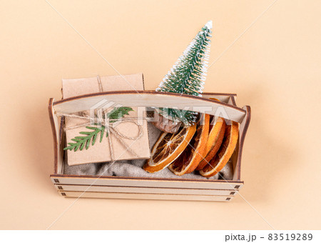 Small wooden box with Christmas decorations - decorative Xmas tree, dry orange slices, DIY wrapped gift box on neutral beige backdrop close up. Holiday image. View from above. Horizontal orientation. Small wooden box with Christmas decorations - decorative Xmas tree, dry orange slices, DIY wrapped gift box on neutral beige backdrop close up. Holiday image. View from above. Horizontal orientation. 83519289
