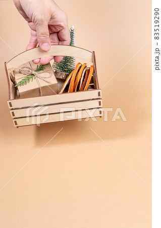 Woman hand holding wooden box with Christmas - decorations decorative Xmas tree, dry orange slices, DIY wrapped gift box on beige background. Christmas image. Vertical orientation. Copy space. Woman hand holding wooden box with Christmas - decorations decorative Xmas tree, dry orange slices, DIY wrapped gift box on beige background. Christmas image. Vertical orientation. Copy space. 83519290