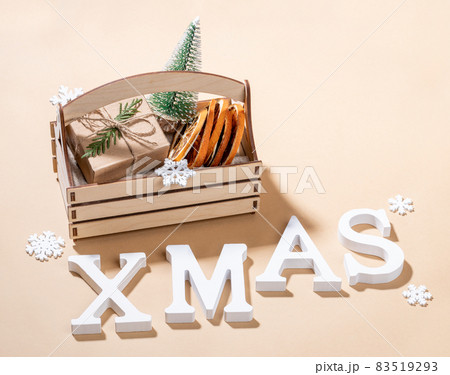 Christmas decorations small decorative Xmas tree, dry orange slices, DIY wrapped gift box in wooden box, snowflakes and white letters XMAS on neutral beige backdrop close up. Festive Christmas image. Christmas decorations small decorative Xmas tree, dry orange slices, DIY wrapped gift box in wooden box, snowflakes and white letters XMAS on neutral beige backdrop close up. Festive Christmas image. 83519293