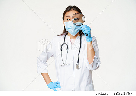 Excited asian medical worker, woman physician looks through magnifying glass surprised, wearing medical face mask and uniform, white background 83521767