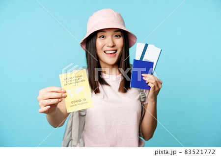 Enthusiastic asian tourist, girl shows covid-19 international vaccination certificate, passport with two tickets, travelling after coronavirus vaccine, blue background 83521772