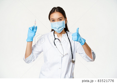Covid-19 vaccination and healthcare. Asian woman doctor in medical face mask and gloves, holding vaccine syringe and show thumb up, white background 83521776