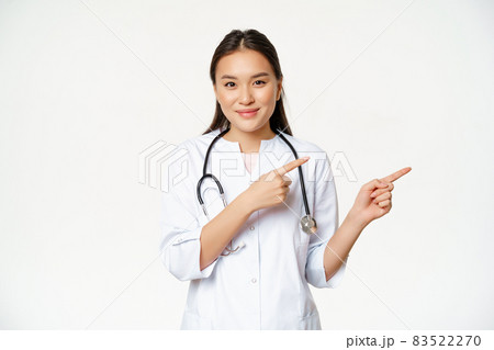 Healthcare, medical people concept. Smiling asian female doctor, pointing fingers right, showing advertisement, hospital info, standing over white background Healthcare, medical people concept. Smiling asian female doctor, pointing fingers right, showing advertisement, hospital info, standing over white background 83522270