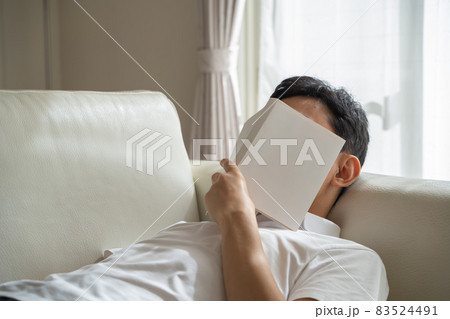 ソファに横になり本を顔にかぶせる男性の様子 83524491