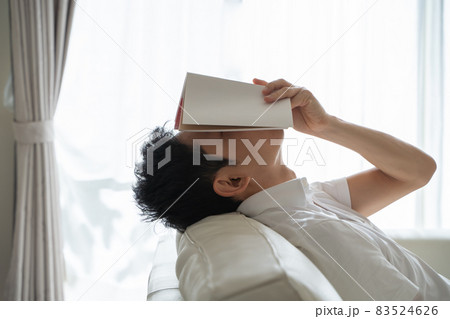 ソファに横になり本を顔にかぶせる男性の様子 83524626