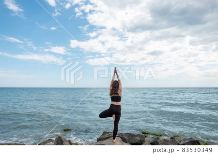 Mudra asana. Greeting woman. Yoga practice. Unrecognizable lady standing in Vrikshasana pose on rock stone namaste hands looking sea view. Mudra asana. Greeting woman. Yoga practice. Unrecognizable lady standing in Vrikshasana pose on rock stone namaste hands looking sea view. 83530349